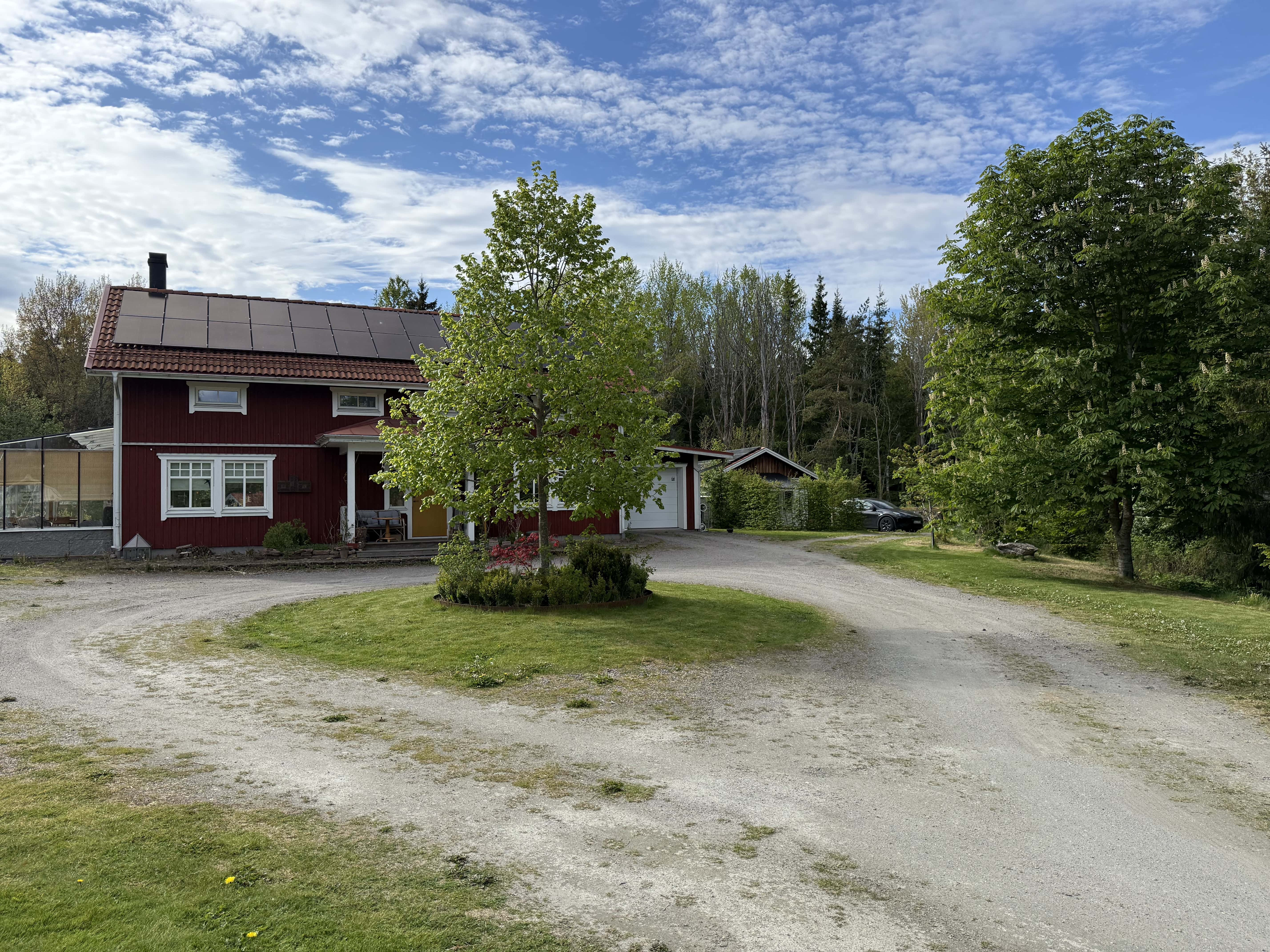 Stenbacken en sommardag — grusväg, gräsmatta och lövträd runt en av stugorna.